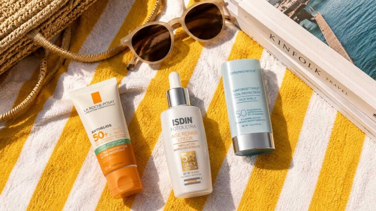 Flat lay of sunscreen products on a yellow striped beach towel with sunglasses, a woven beach bag, and a travel book under natural sunlight.